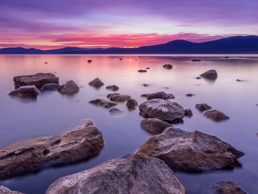 Lake Tahoe at sunset, with smooth granite boulders in calm water and mountains under a pink and violet sky